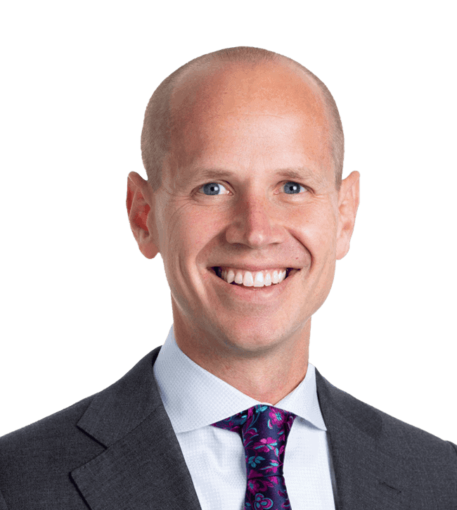 Professional headshot: smiling bald man in a dark suit and purple patterned tie, facing the camera against a green background.