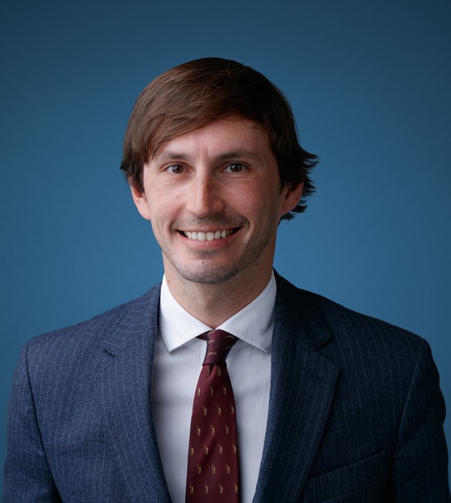 Smiling man in a navy pinstripe suit with white shirt and burgundy tie, posed against a blue backdrop.